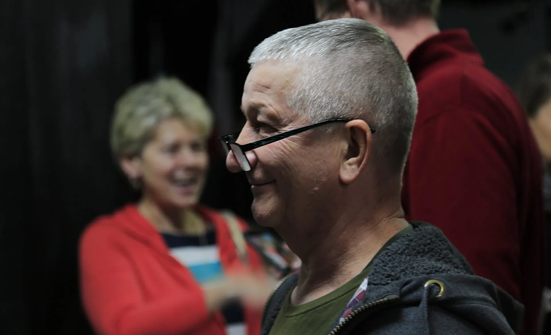 A smiling older man in glasses, a laughing woman in a red sweater behind him.