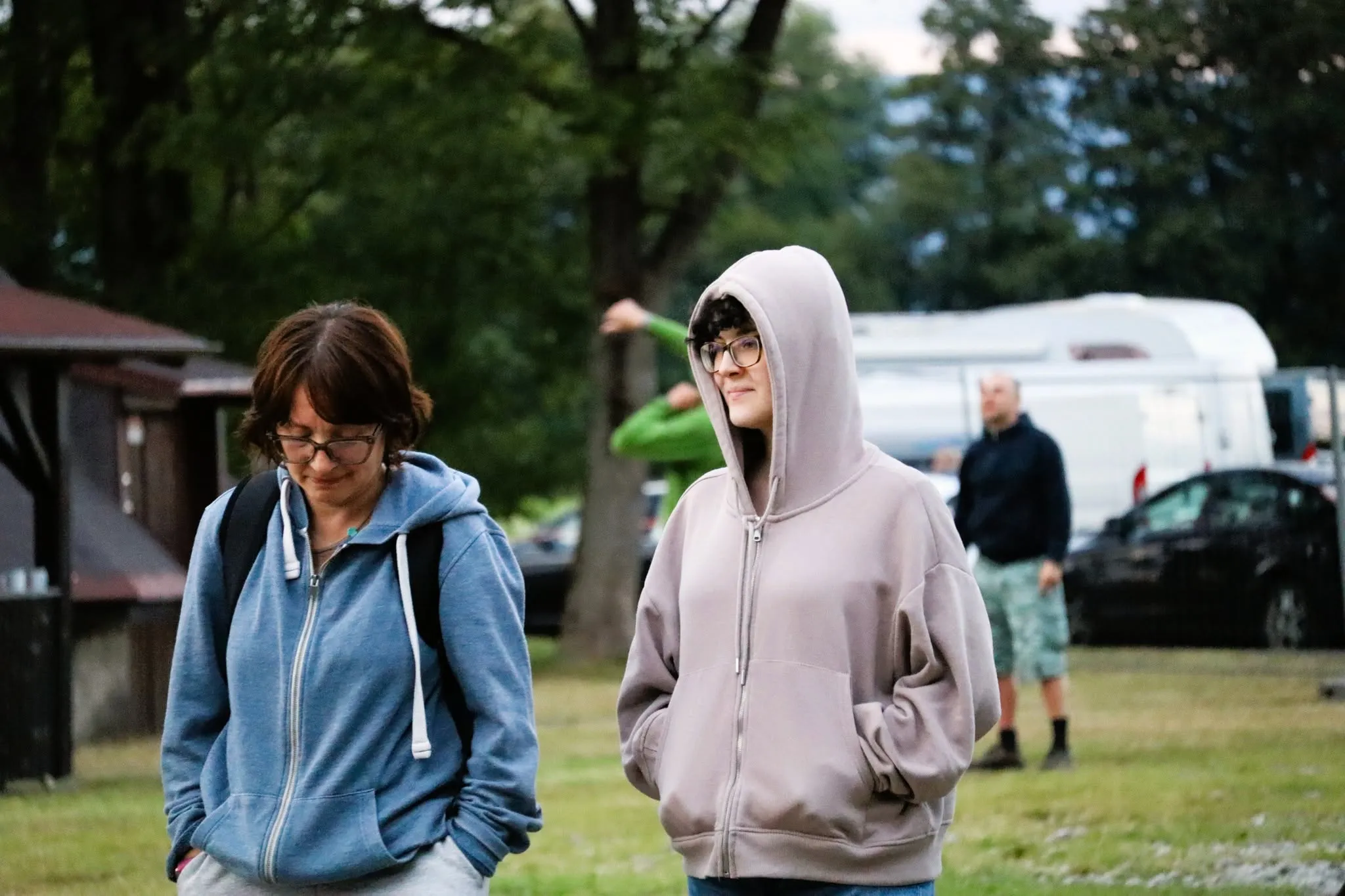 Two people in hoodies walking through a campground; a camper and trees behind them.