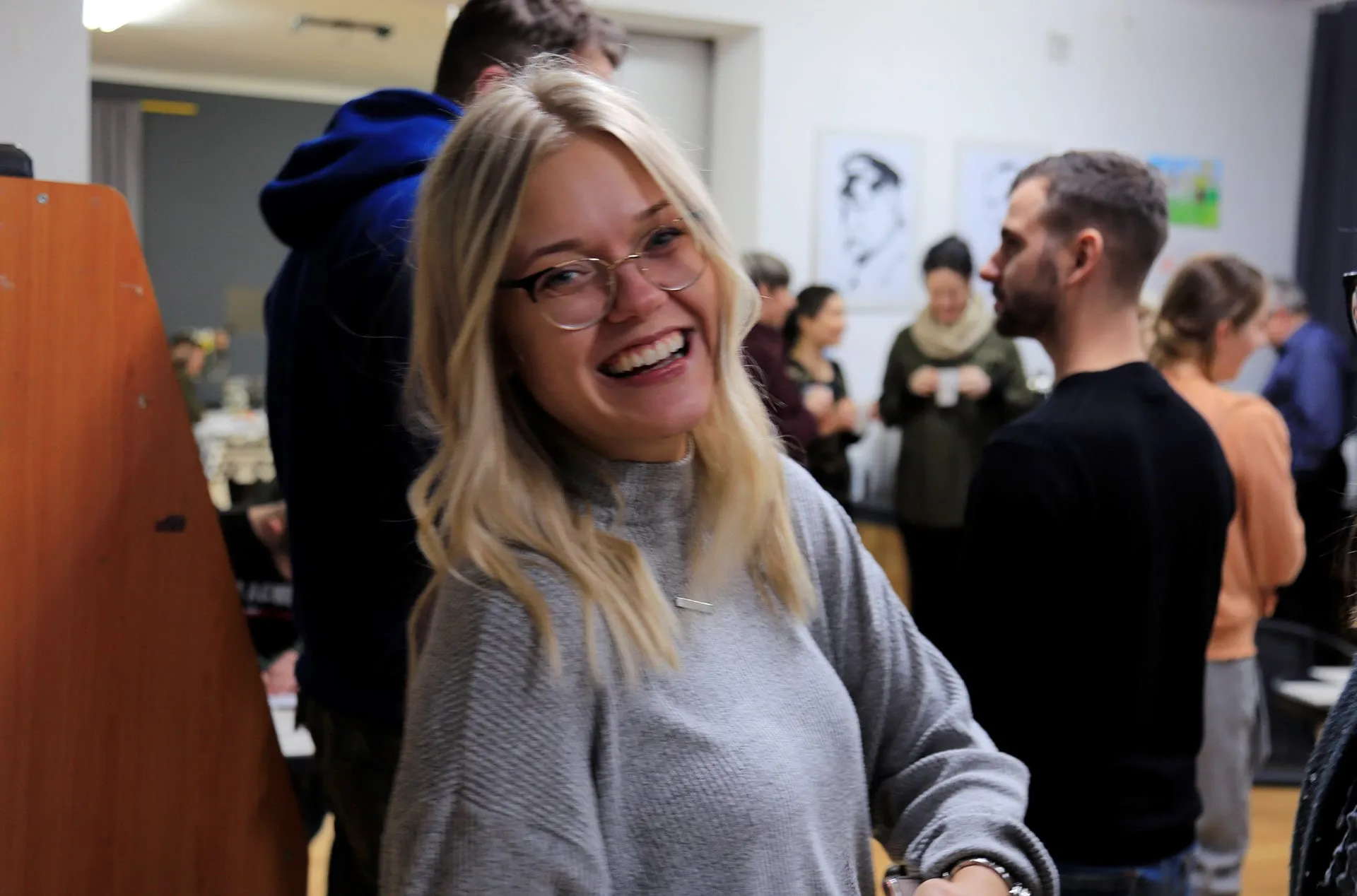 A smiling young woman in glasses, conversations in the background.